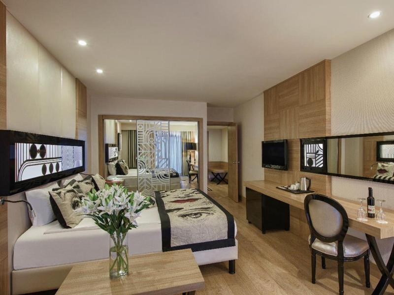 Moderní hotelový pokoj s postelí, květinami, psacím stolem, židlí a velkým zrcadlem.