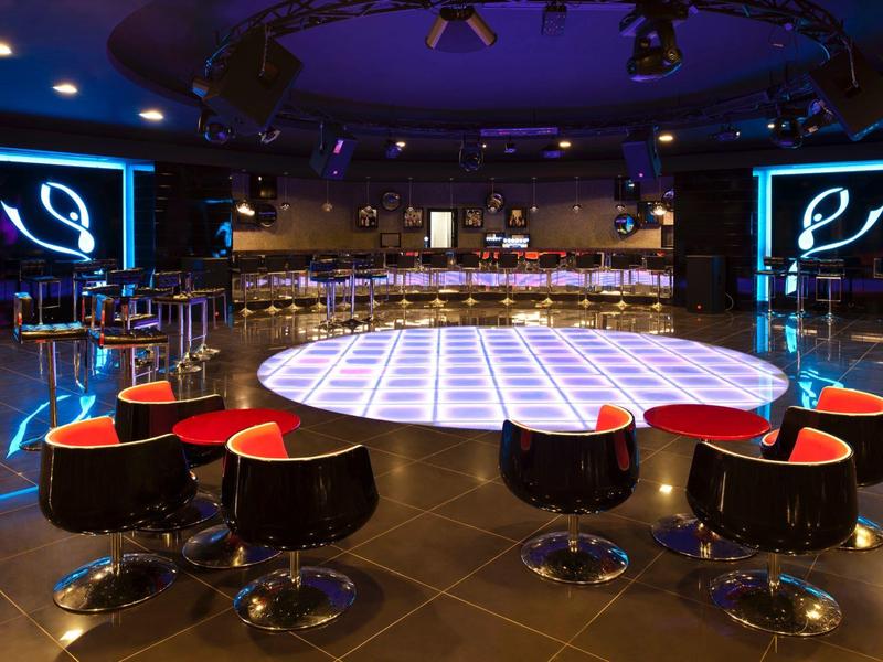 Moderní bar s kruhovým sezením kolem jasně osvětleného tanečního parketu pod modrými reflektory.