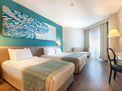 Helles Hotelzimmer mit zwei Doppelbetten, türkisfarbener Wand und großem Fenster mit Vorhängen.