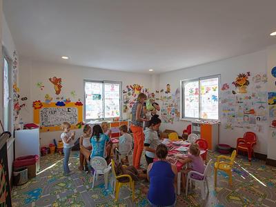 Kindergartenraum mit Kindern, bunten Stühlen, Wandzeichnungen und hellem Tageslicht.