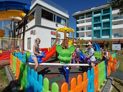 Kinder spielen auf einem bunten Karussell in einem umzäunten Spielplatz vor Wohngebäuden.
