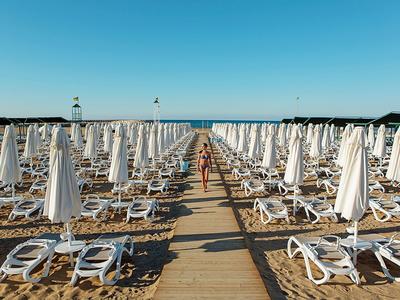 Strand mit Reihen von weißen Sonnenschirmen und Liegen, Holzsteg führt zur Wasserlinie.