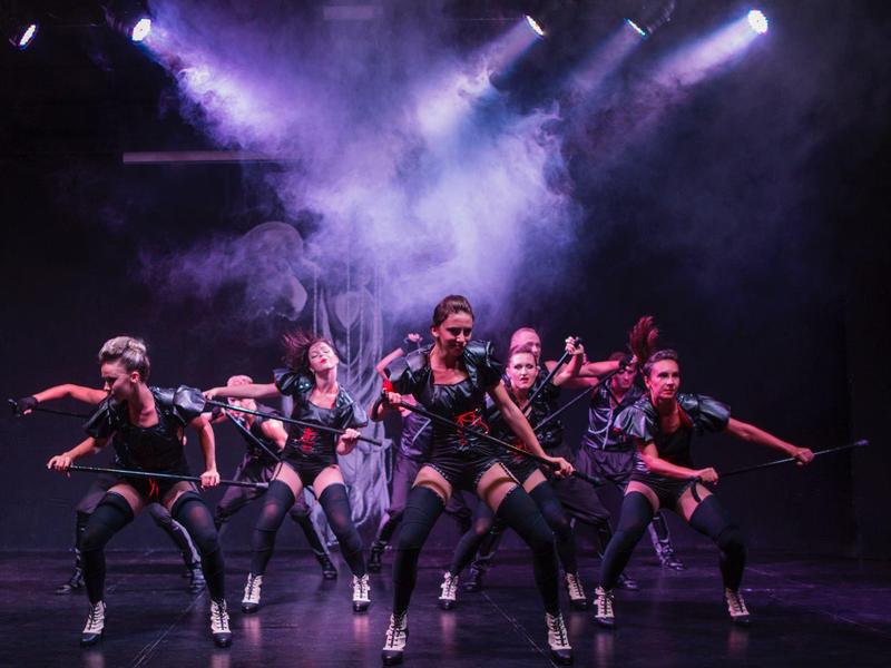 Een groep dansers voert een energieke routine uit op een donker podium met rookeffecten.