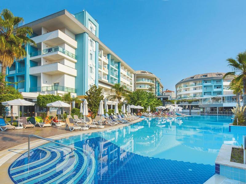 Modern hotelcomplex met groot zwembad, ligstoelen en palmbomen bij zonnig weer.