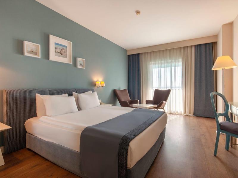 Moderne hotelkamer met tweepersoonsbed, stoelen en bureau bij een groot raam.