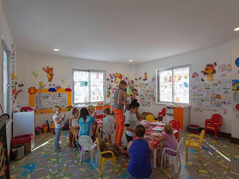 Een lichte kamer met kinderen en volwassenen die spelen aan kleurrijke tafels en stoelen, met wanddecoraties.