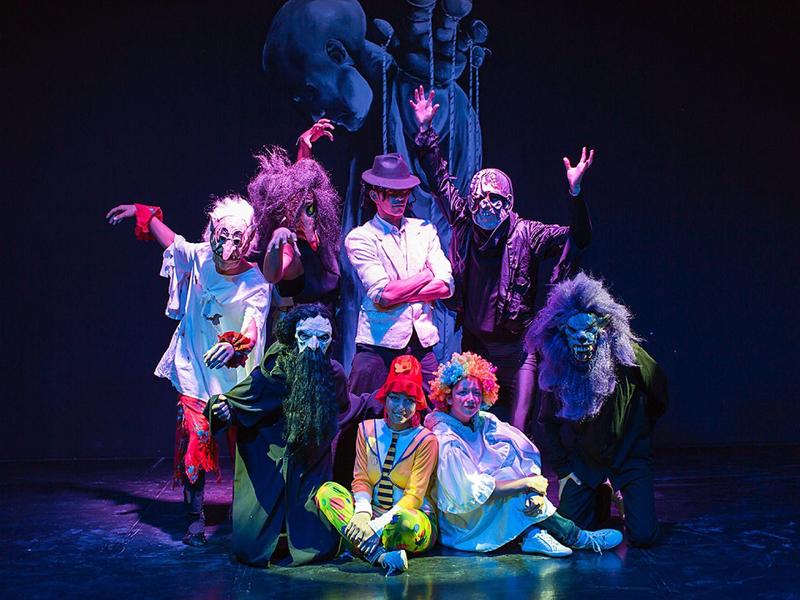 Zes mensen in kostuums en make-up poseren op een podium met een donkere achtergrond.