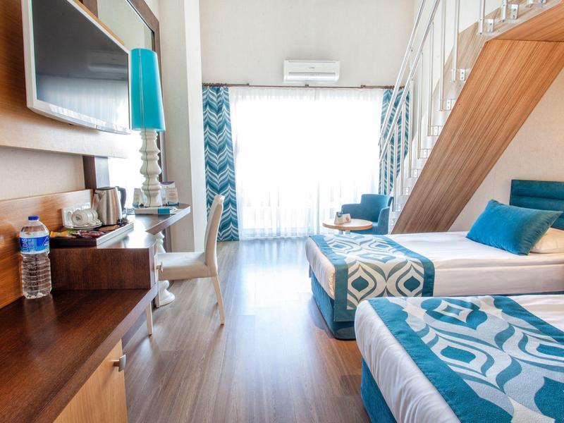 Chambre d'hôtel lumineuse avec deux lits, accents bleus et grande fenêtre avec rideaux.