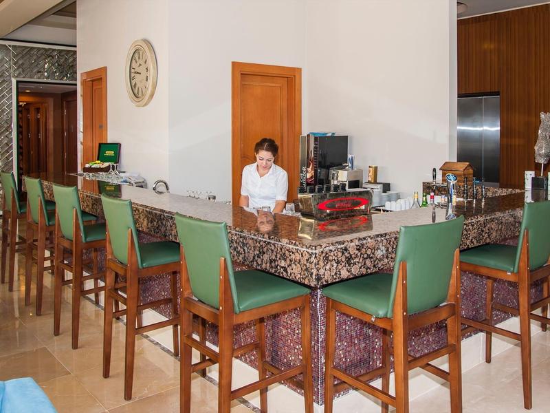 Comptoir avec tabourets en bois verts rembourrés et barmaid en blouse claire dans un hall d'hôtel élégant.