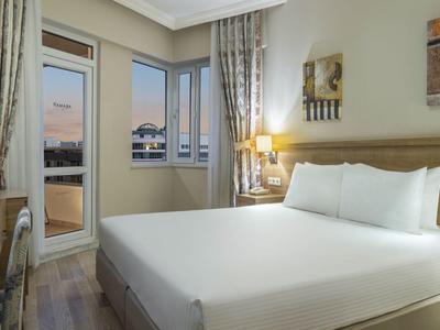 Camera d'hotel moderna con letto matrimoniale, scrivania, TV e vista sulla città al tramonto.