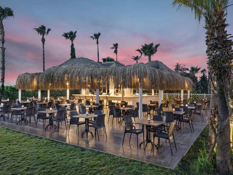 Terrazza con tavoli sotto tettoie di paglia illuminate al crepuscolo in ambiente tropicale.