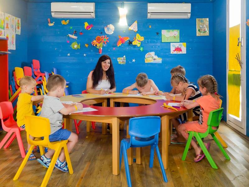 Children sit around a table in a colorful room with blue walls, engaging in crafts.