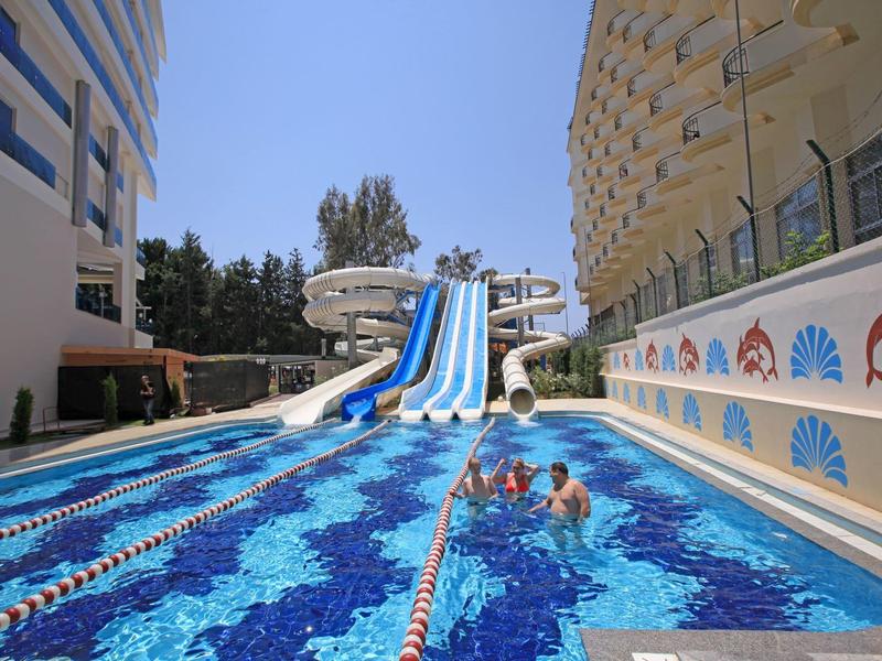 Venkovní bazén s vodními skluzavkami mezi dvěma hotelovými budovami za slunečného dne.