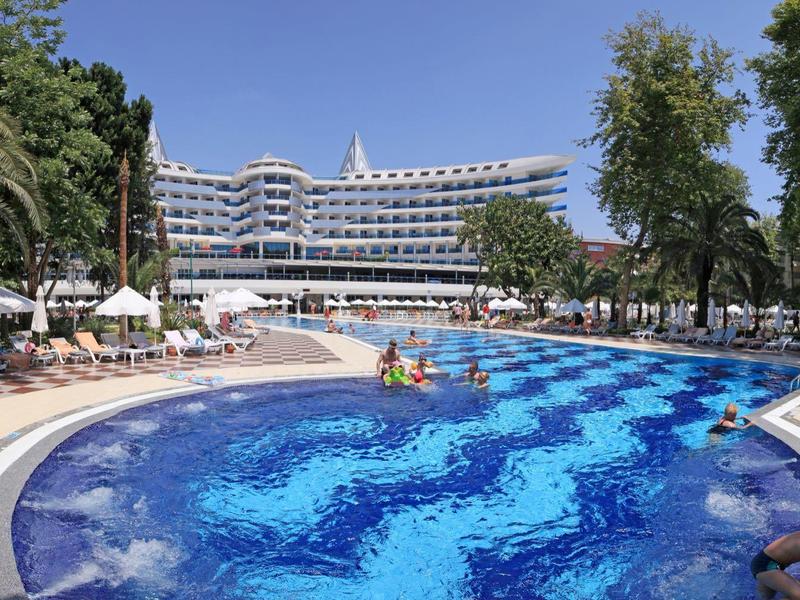 Velký bazén s vlnitým vzorem před vícepodlažním hotelem obklopeným palmami.