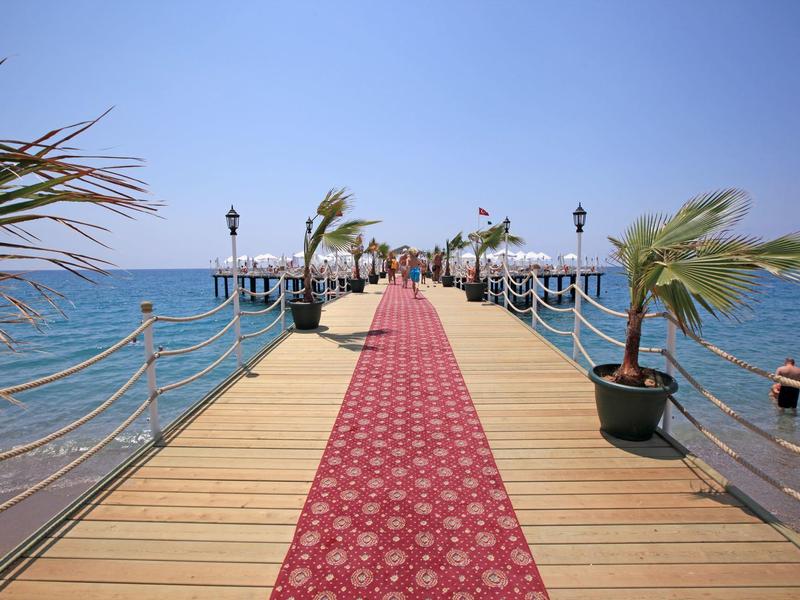 Molo s červeným kobercem, palmami a lampami vedoucí do moře pod jasnou oblohou.