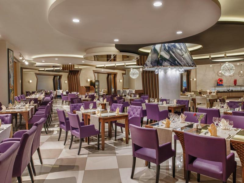 Velká hotelová jídelna s mnoha stoly a fialovými židlemi pod nepřímým osvětlením.