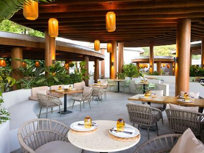 Restaurant ouvert avec tables rondes, chaises et lampes suspendues dans un cadre tropical.