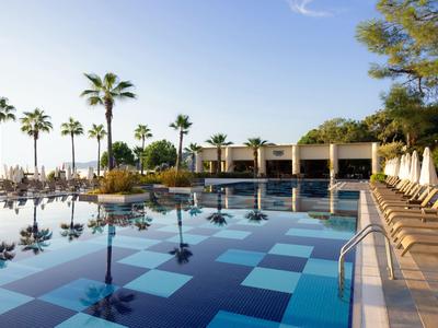 Piscine d'hôtel moderne avec palmiers, parasols et chaises longues sous un ciel bleu.