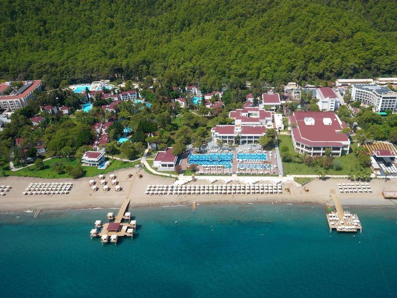 Vue aérienne d'une station balnéaire avec plage, piscines et bâtiments entourés de verdure.