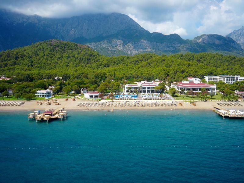 Plage avec eau turquoise, structures en bois, et montagnes en arrière-plan sous un ciel partiellement nuageux.