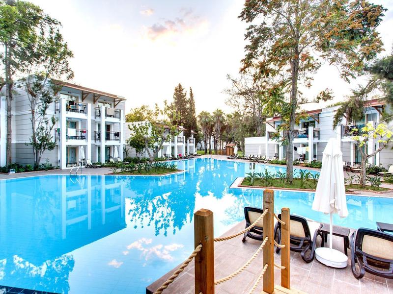 Piscine bleue entourée d'arbres et de bâtiments blancs avec terrasses et chaises longues.