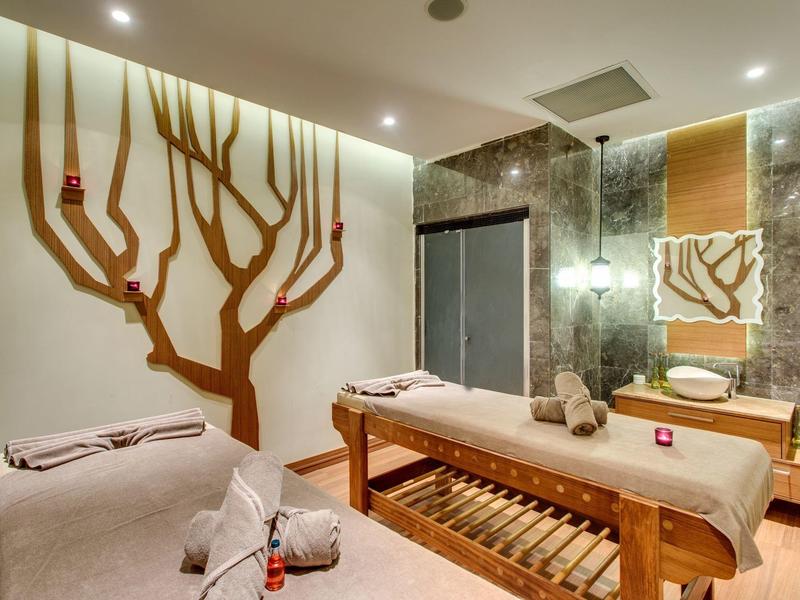 Salle de massage chaleureuse avec tables, décoration murale en forme d'arbre, éclairage doux.