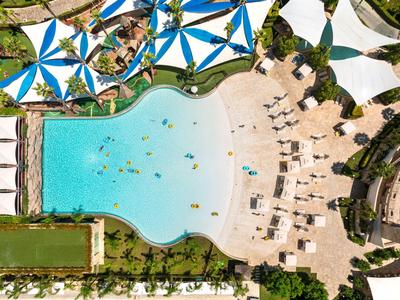 Luchtfoto van een resortzwembad met blauw-witte parasols en ligbedden rondom het water.