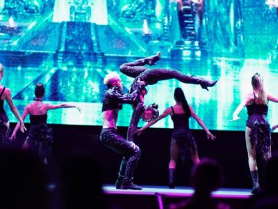 Dancers perform an acrobatic lift on stage with a colorful LED background wall.