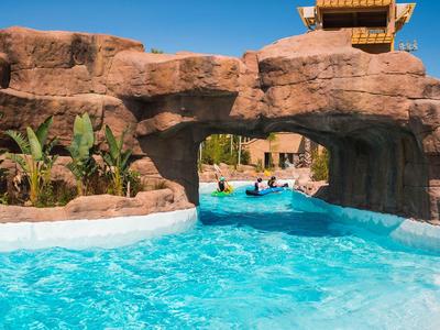 Buitenzwembad met helder blauw water dat onder een rotsboog doorstroomt, omgeven door palmbomen.