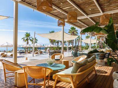 Offener Strand-Lounge-Bereich mit Holzmöbeln, Palmen und Blick auf Meer und Strandliegen.