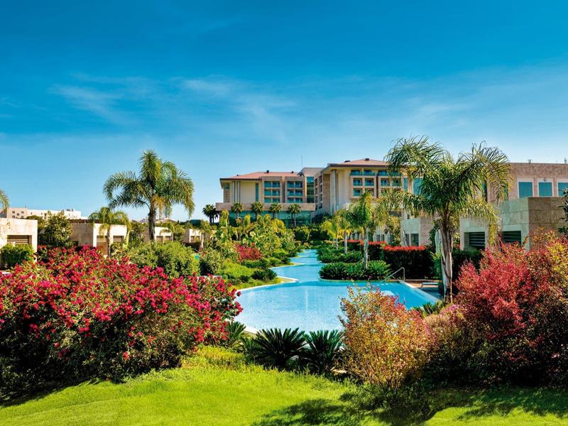 Luxe hotel met zwembad, omgeven door tropische planten en palmbomen onder een heldere blauwe lucht.