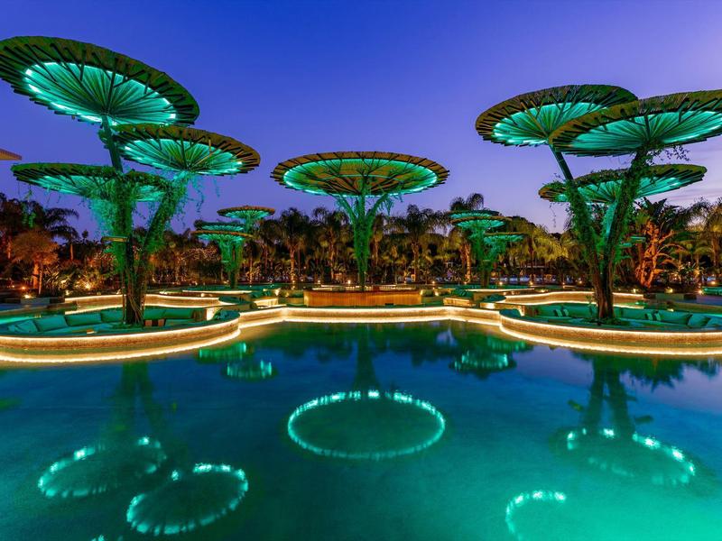 Futuristische bomen met groene verlichting weerspiegeld in een hotelzwembad bij schemering.