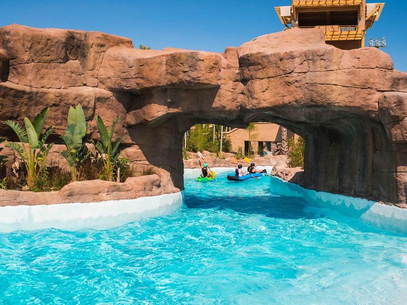 Buitenzwembad met helder blauw water dat onder een rotsboog doorstroomt, omgeven door palmbomen.