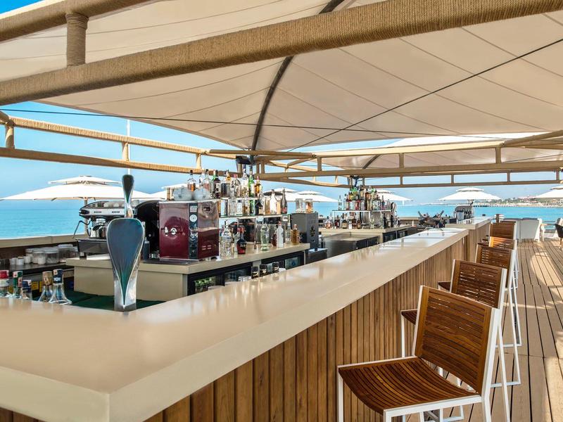 Buitenbar met houten krukken onder een luifel, met uitzicht op zee op een zonnige dag.