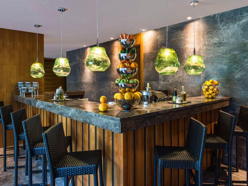 Gezellige hotelbar met houten toonbank, zwarte krukken, groene hanglampen en decoratieve fruitkommen.