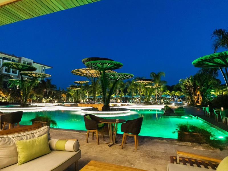 Luxe zwembad bij nacht met verlicht water, loungeset en tropisch groen.