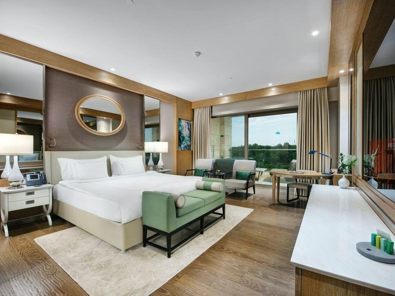 Ruime hotelkamer met groot bed, zithoek, houten vloeren en groot raam met balkonuitzicht.