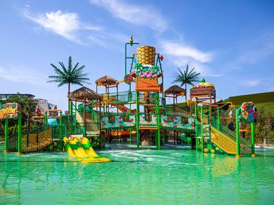 Parco acquatico colorato con scivoli, spruzzi e palme sotto un cielo blu.