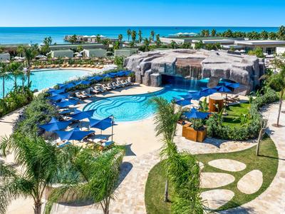 Grande piscina all'aperto con sedie a sdraio blu, palme e vista sul mare in un resort.