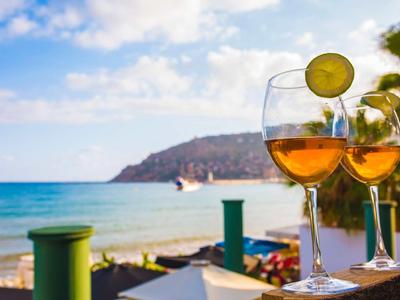 Two wine glasses with lime slices on a beach with sea and hills in the background.