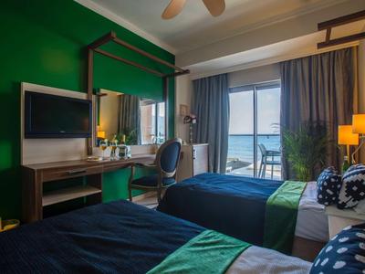 Camera d'hotel con due letti, parete verde e balcone con vista mare.