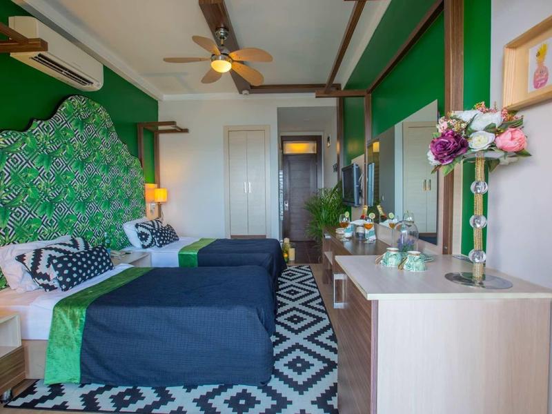 Camera d'albergo moderna con due letti, accenti verdi, ventilatore a soffitto e composizione floreale.