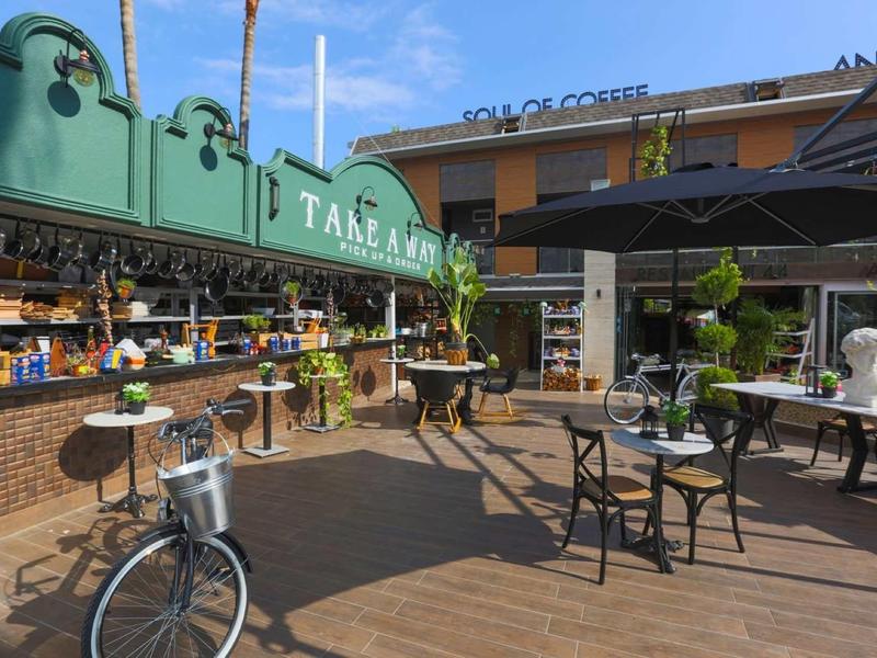 Area esterna di un caffè con tavoli, sedie e un portamanubrio per bici con tempo soleggiato.