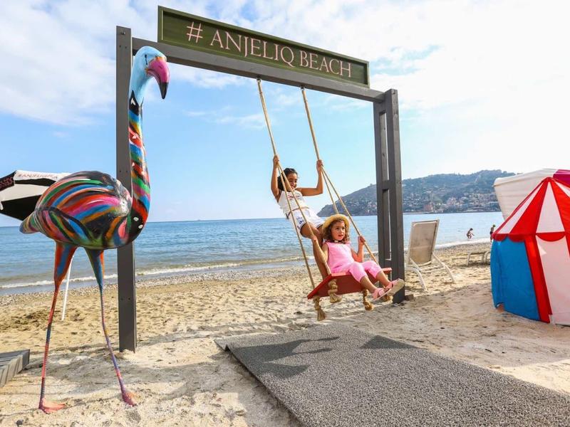 Due bambini su un'altalena sulla spiaggia con una scultura di fenicottero colorata e una tenda sullo sfondo.