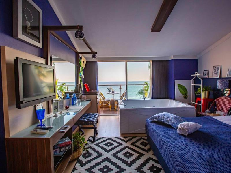 Camera moderna con letto blu, scrivania, tappeto e vista mare attraverso grandi finestre.