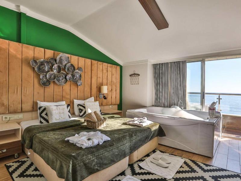 Camera d'hotel luminosa con parete in legno, letto matrimoniale, divano e grande finestra con vista mare.