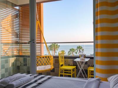 Balkon mit gelben Stühlen und Tisch, Blick aufs Meer, Raum mit gestreiften Vorhängen und Bett.