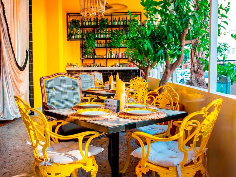 Bunte Café-Terrasse mit gelben Stühlen, Holztisch, grünem Baum und Regal voller Flaschen.