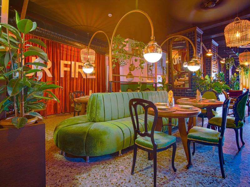 Modern eingerichtetes Café mit grünem Sofa, Holztischen, Pflanzen und hängenden Lampen.