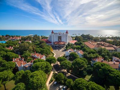 Pohled na velký hotel u moře, obklopený zelenými stromy a malými domy.